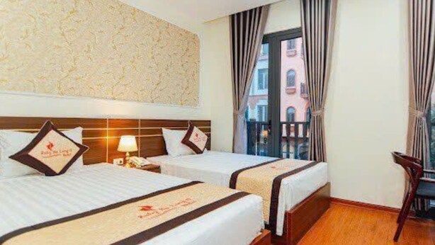 A hotel room with two beds, a wooden headboard, a nightstand, a lamp, and a view of buildings through a window with curtains. -  Ruby Ha Long 6 Hotel (Bai Chay)