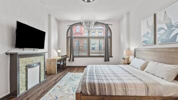 A bedroom with a large bed, a flat-screen TV, a fireplace, and a window with blinds.