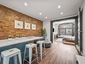Una cocina moderna con pared de ladrillo, taburetes de bar blancos y una zona de comedor con sofá gris.