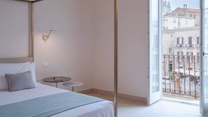 A hotel room with a bed, a small table, a wall-mounted light, and a balcony with a view of buildings. - Palazzo Pupillo Liberty Suite (Syracuse)