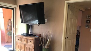 A living room with a television on a wooden cabinet, a green vase with decorative branches, a patterned rug, and a door leading to another room. - Available Now! -Utilities Included-Prime Spot!Pools- Spas-shopping-restaurant’s  (Phoenix)