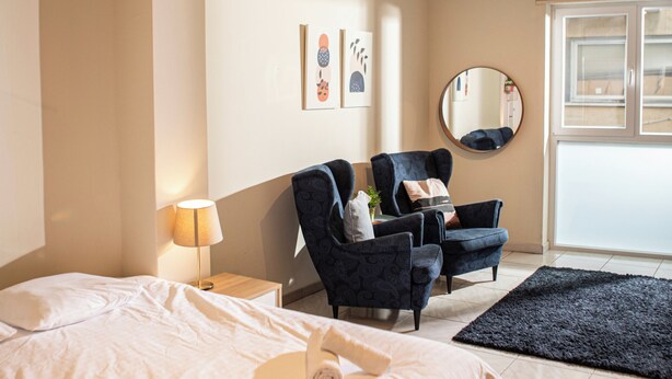 A hotel room with a large bed, two armchairs, a small table, a lamp, a round mirror, and artwork on the wall. - Charming studio with reading nook & city view (Nicosia)