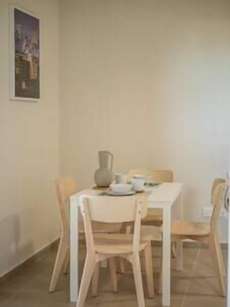 A dining area with a white table and four wooden chairs. There is a vase on the table and a framed picture on the wall.