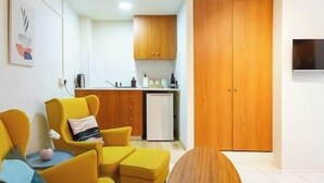 Een knus woongedeelte met een kitchenette, een houten garderobe en een gele fauteuil met een klein tafeltje.