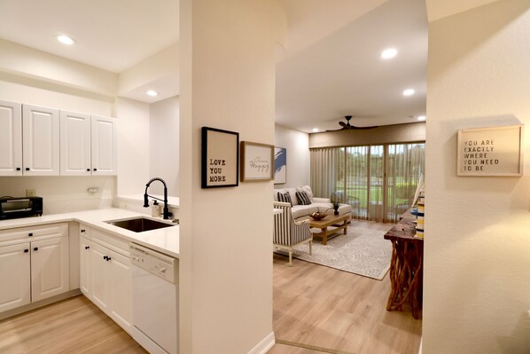 A modern kitchen with white cabinetry, a built-in dishwasher, and a sink. There is a dining area with a striped chair and a table, and a living room with a sofa and a coffee table.