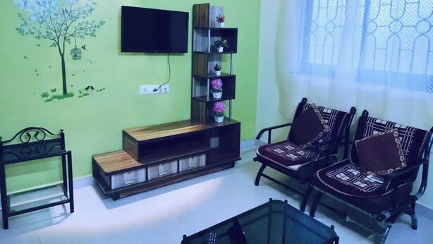 Un salon doté d’un téléviseur à écran plat fixé au mur, d’un meuble de rangement en bois, de deux fauteuils à motifs, d’une petite table et d’une fenêtre ornée de rideaux légers.