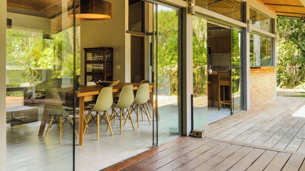 Un espace de restauration extérieur moderne, doté d’une terrasse en bois, de murs en verre et offrant une vue sur la verdure.
