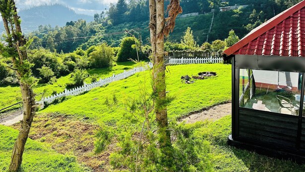 A view of a lush green landscape with a white picket fence, a small building with a red roof, and a tree in the foreground. - Hoopoe House (Ootacamund)