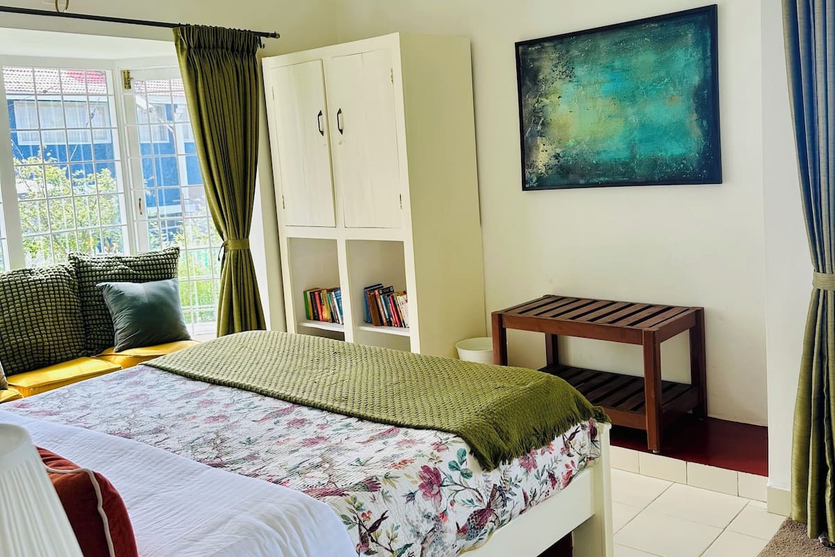 A bedroom with a bed, a white wardrobe, a small table, a bookshelf, and a painting on the wall.