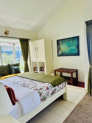A bedroom with a bed, a white wardrobe, a small table, a bookshelf, and a painting on the wall. - Hoopoe House (Ootacamund)