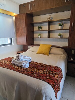 Une chambre à coucher avec un lit, un oreiller jaune, une serviette enroulée, une couverture à motifs et des armoires en bois avec des étagères au-dessus du lit.
