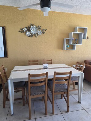Un coin repas avec une table blanche, des chaises en bois, un ventilateur de plafond et des œuvres d’art décoratives sur les murs.