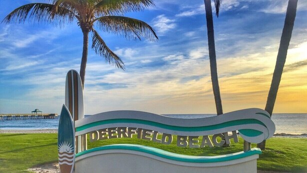 A sign for Deerfield Beach with a surfboard design, palm trees, and a view of the ocean.