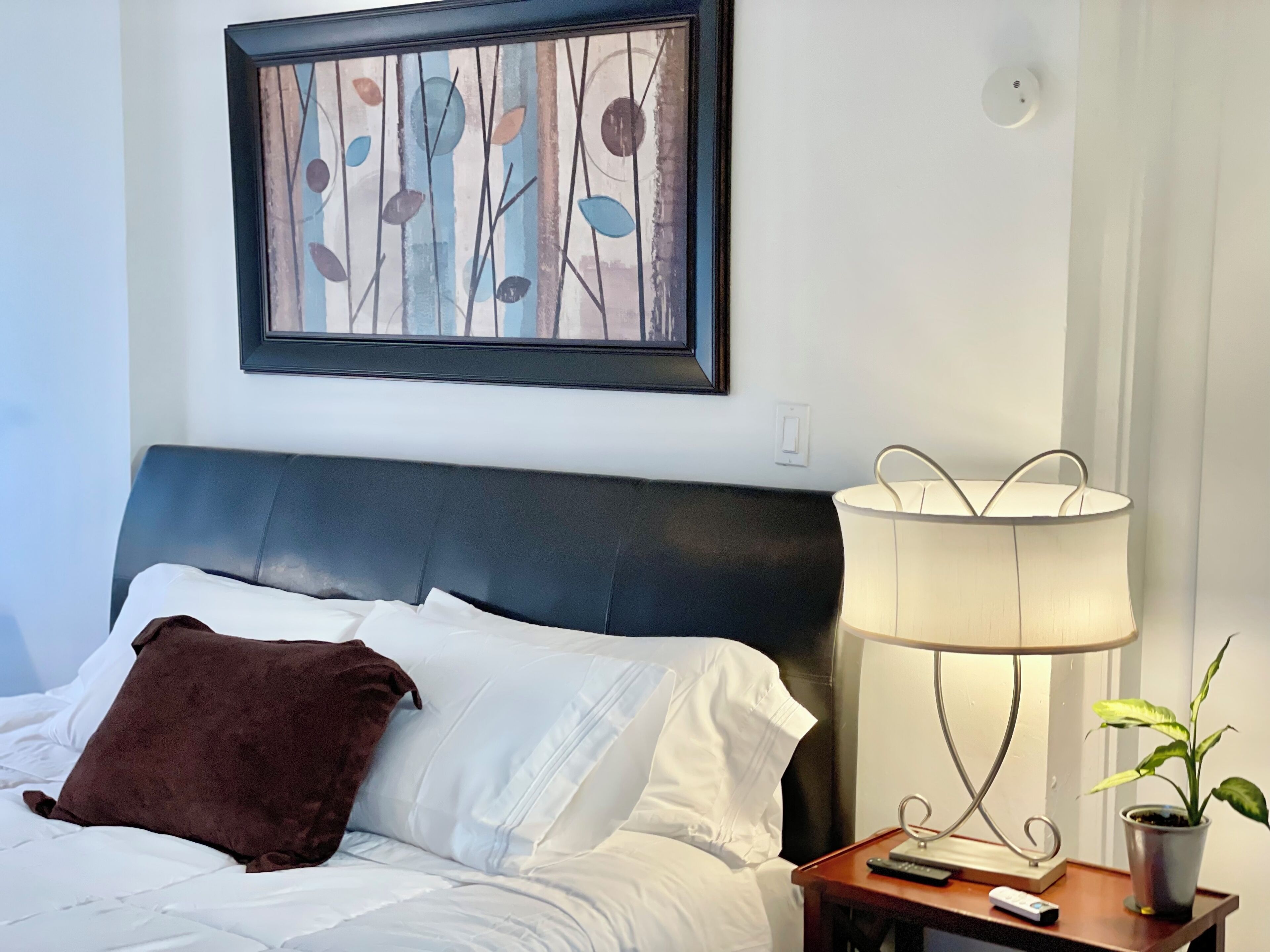 Cama bem arrumada com lençóis brancos e travesseiro marrom, mesa de cabeceira com abajur e controle remoto, e pintura abstrata emoldurada na parede.