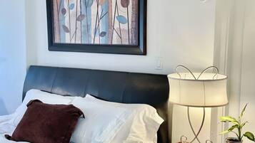 Cama bem arrumada com lençóis brancos e travesseiro marrom, mesa de cabeceira com abajur e controle remoto, e pintura abstrata emoldurada na parede.