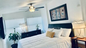 A bedroom with a bed, a ceiling fan, a nightstand, a lamp, a potted plant, and a painting on the wall.