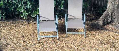 Duas cadeiras de metal com assentos e encostos de tecido, posicionadas sobre um piso de cascalho em uma área de jardim com cerca de madeira e folhagem ao fundo.