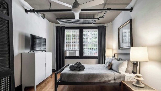 A bedroom with a bed, a ceiling fan, a TV, a window with blinds, a bedside table with a lamp, and a wardrobe. - The Burrow at Wrigley (Chicago)