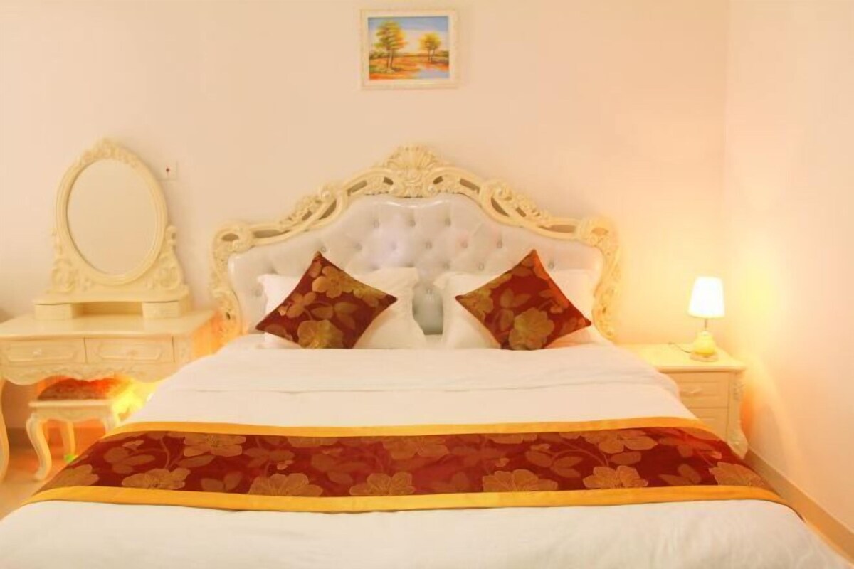 Quarto com cabeceira estofada, duas almofadas, mesa de cabeceira com abajur e um pequeno quadro na parede.