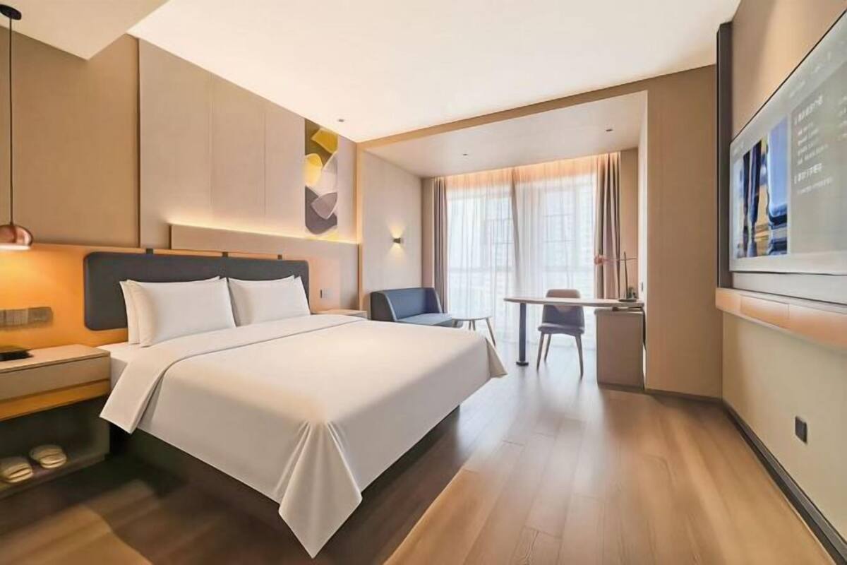 A hotel room with a large bed, bedside table, lamp, and a desk with a chair.