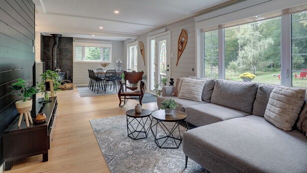 A modern living room with a sectional sofa, a dining area with a table and chairs, and large windows offering a view of the outdoors. - Hôtel à la maison - La Maison Rouge (Potton)