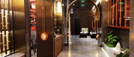 A corridor with a modern interior design, featuring a central light fixture, a seating area, and a decorative plant arrangement.