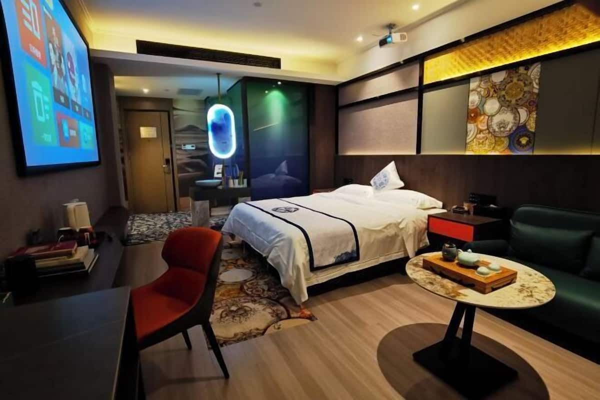 A hotel room with a bed, a desk, a chair, a small table, and a TV screen.