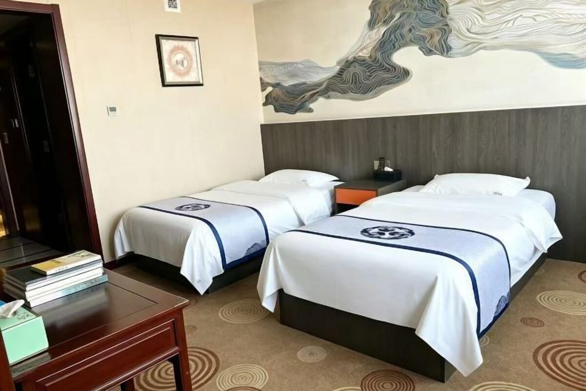 Een hotelkamer met twee bedden, een houten nachtkastje, een bureau met boeken en een groot abstract schilderij aan de muur.
