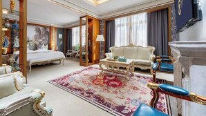 A luxurious hotel room with a large bed, a sitting area with a sofa and armchairs, a fireplace, and a decorative rug.
