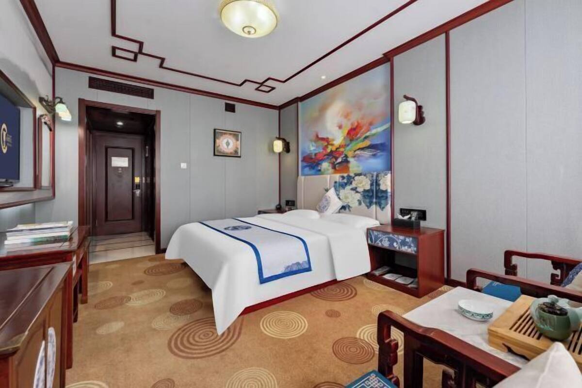 A hotel room with a bed, bedside table, and a desk with a chair. There is a painting on the wall and a door leading to another room.