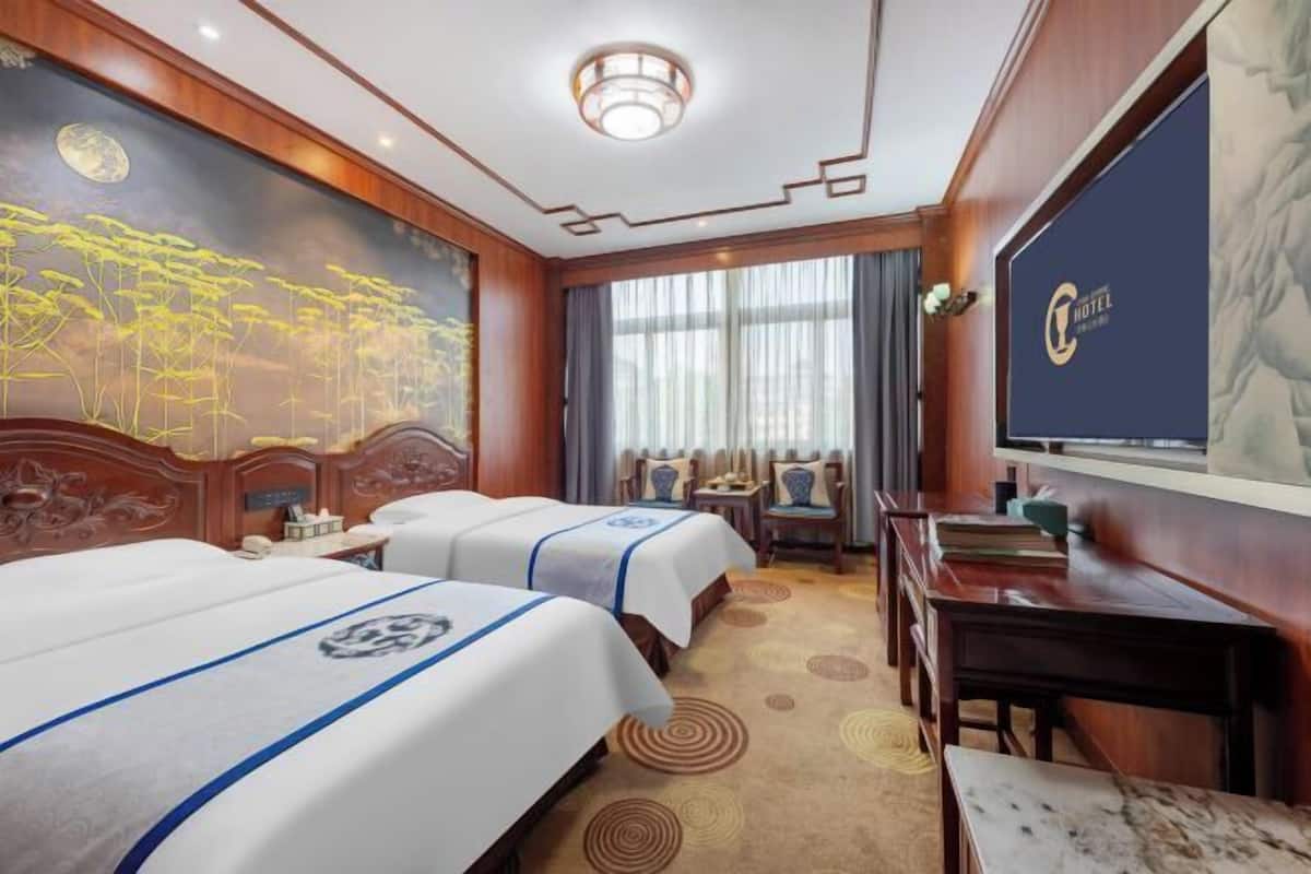 A hotel room with two beds, a desk, and a large screen displaying the hotel name.
