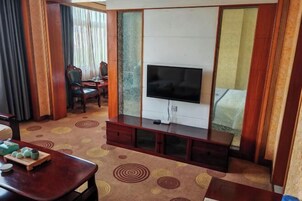 Habitación de hotel con televisor, un mueble de madera, una mesa de comedor con juego de té y una alfombra estampada.