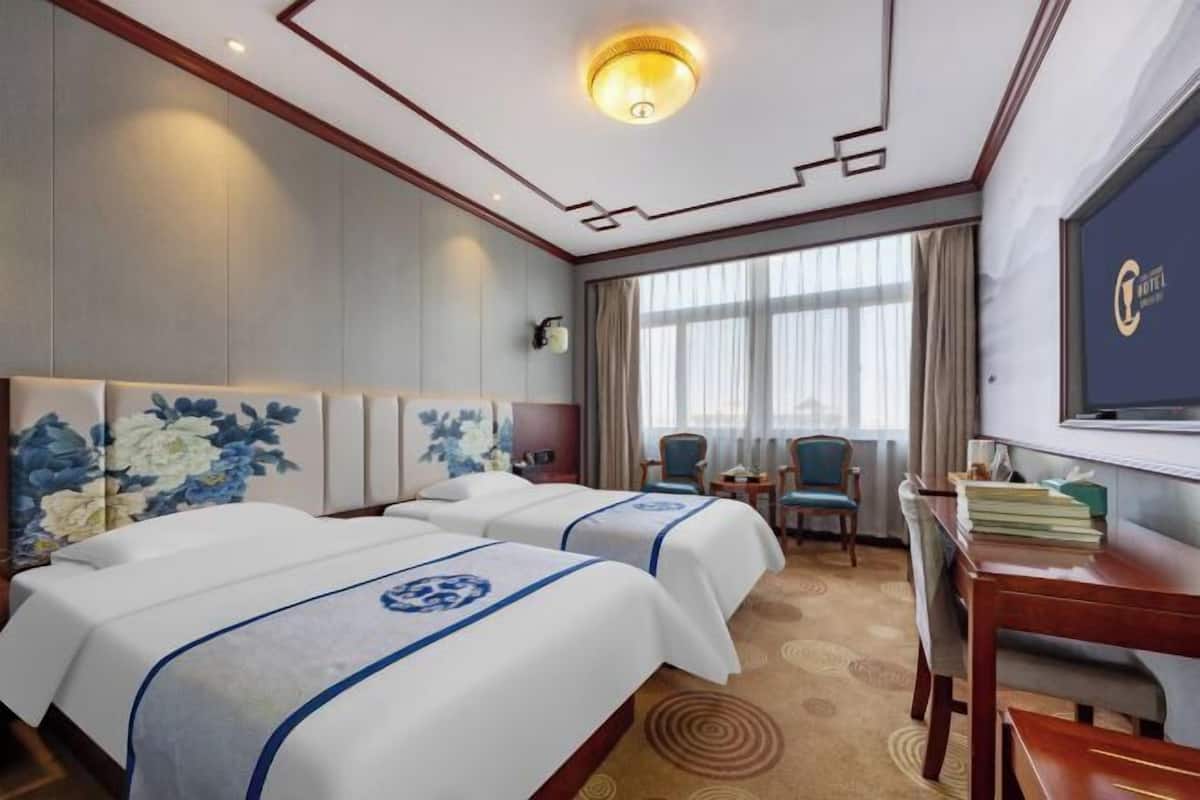 A hotel room with two beds, a desk, a chair, a TV, and a large window with curtains.