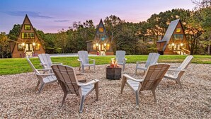 A cozy outdoor seating area with Adirondack chairs and a fire pit, surrounded by a gravel ground, with a backdrop of wooden cabins and a twilight sky. - Adults-Only Luxury Cabin w/ Steam Shower + HotTub (Omaha)