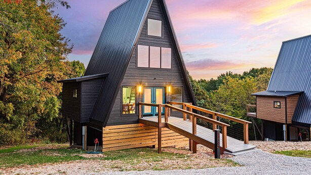 A modern, triangular house with a wooden deck and a staircase leading to the entrance.