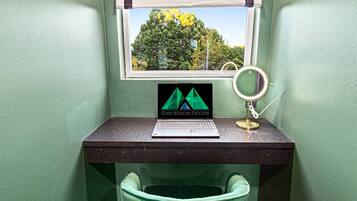 Un petit bureau avec un ordinateur portable, un anneau lumineux et une poubelle, dans une pièce aux murs verts et dotée d’une fenêtre.