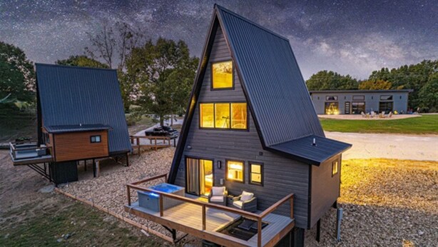 Una cabaña moderna de forma triangular con terraza y zona de estar exterior, rodeada por un paisaje de grava y un cielo estrellado.
