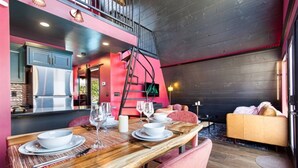 Comedor con mesa de madera dispuesta para cuatro personas, una escalera y un sofá al fondo.