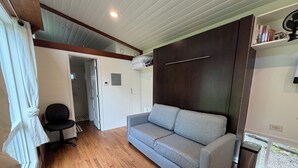 A room with a grey sofa, a black office chair, wooden flooring, a ceiling fan, and a window with curtains. - Charming Tiny House Retreat (Olympia)