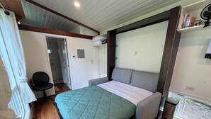 A bedroom with a bed, a chair, a small table, a window, and a door. - Charming Tiny House Retreat (Olympia)