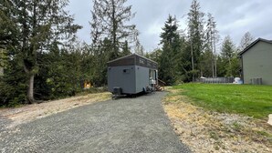 A small, gray mobile home with a gravel driveway, surrounded by dense trees and a grassy area. - Charming Tiny House Retreat (Olympia)