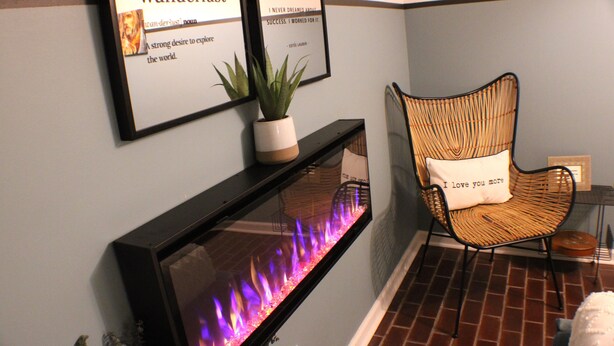 A fireplace with a fire, a wicker chair with a cushion, and two framed quotes on the wall.