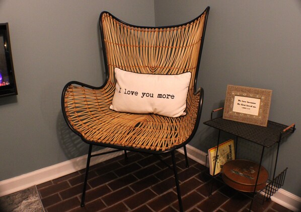 A wicker chair with a cushion that reads "I love you more." Next to the chair is a small table with a framed sign and a picture frame.