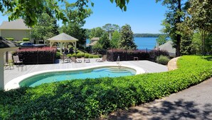 A swimming pool with a curved edge, surrounded by a deck and lounge chairs, with a gazebo and a view of a lake.