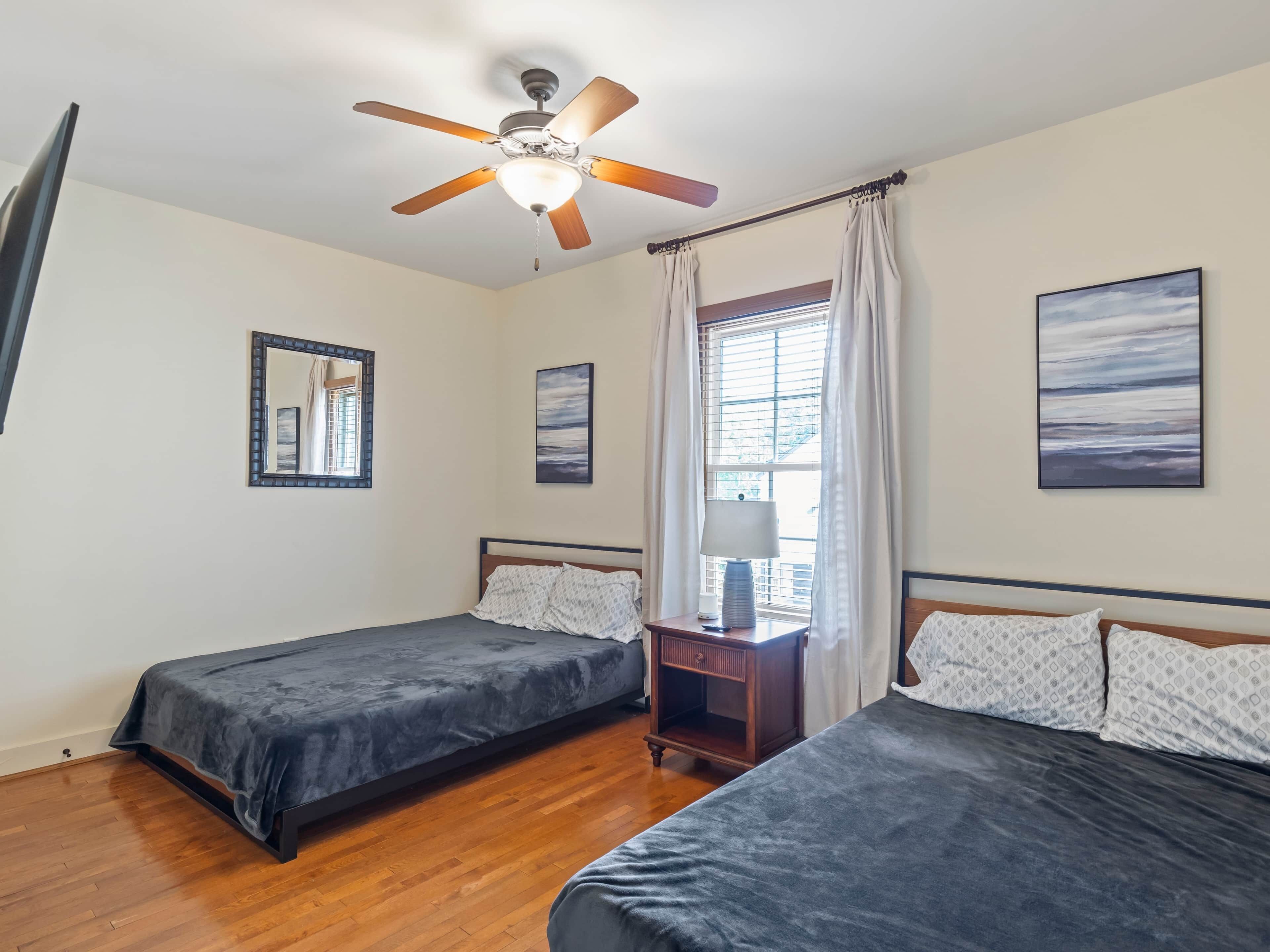 Quarto com duas camas, um ventilador de teto, uma janela com cortinas e duas imagens penduradas na parede.
