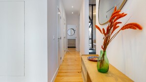 Een hal met houten vloeren, een houten bankje, een groene vaas met gedroogde planten en een ronde spiegel aan de muur.
