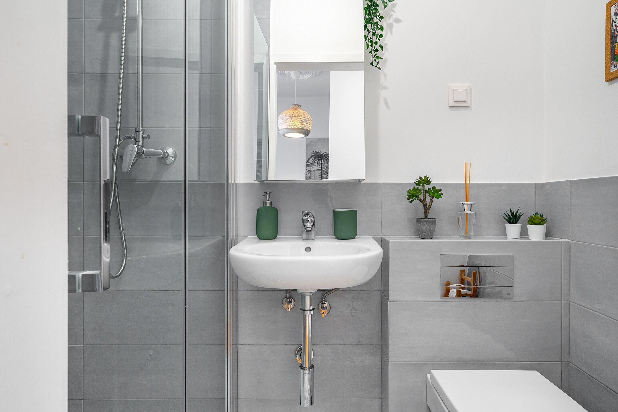 Une salle de bain moderne avec une douche à l’italienne en verre, un lavabo blanc agrémenté de tasses vertes et une petite étagère avec des plantes et des articles de toilette.