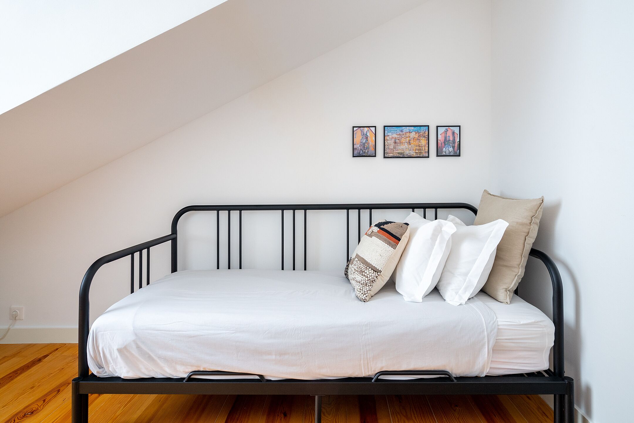 A bed with a white mattress, black metal frame, and multiple pillows. There are three framed artworks on the wall above the bed.