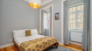 Un dormitorio con cama, colcha estampada, lámpara colgante, ventana con cortinas y radiador.