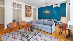 A modern living room with a blue accent wall, a grey sofa, a glass coffee table, and a dining area with a table and chairs. - Alecrim 43 Top Location Perfect To Explore Lisbon (Lisbon)